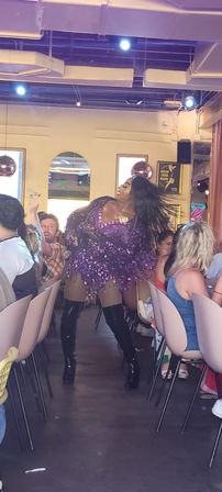 Drag performer in a purple sequined fringe dress and black thigh-high boots struts through a crowded restaurant during a lively brunch show.
