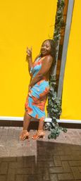 Smiling woman in a blue-and-orange tropical dress and orange high heels posing with a peace sign against a bright yellow wall with green vines on a brick sidewalk — sunny outdoor scene.