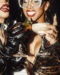Glamorous nightlife shot of two people toasting with coupe cocktails — one mint-garnished, spiced-rim drink held in a glossy black latex glove and a frothy cocktail in manicured fingers, surrounded by sequined fashion, rhinestone sunglasses and bold red lipstick.