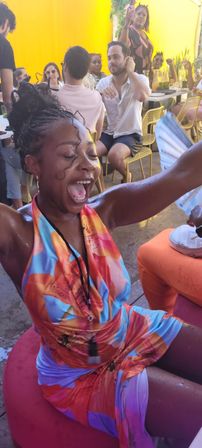 Joyful woman in a colorful floral halter dress with arms raised, laughing and dancing at a lively outdoor patio brunch with friends under a sunny yellow backdrop