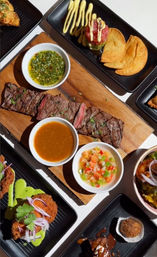 Overhead shot of sliced grilled steak on a wooden board with chimichurri, brown dipping sauce and fresh pico de gallo, surrounded by crispy plantain chips, fried croquettes and assorted appetizers on black plates