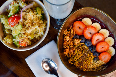 Top-down view of vibrant brunch: an acai bowl topped with sliced strawberries, banana, blueberries, granola, pumpkin and hemp seeds, beside a quinoa salad with cucumber, cherry tomatoes and red onion on a wooden table with a spoon and napkin.