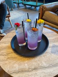 Four purple ombré drinks in tall glasses with black straws and colorful edible flower garnishes, served on a black tray atop a marble cafe table in a cozy lounge setting.