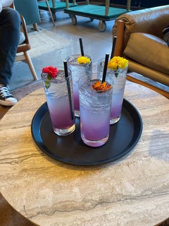 Four purple ombré drinks in tall glasses with black straws and colorful edible flower garnishes, served on a black tray atop a marble cafe table in a cozy lounge setting.