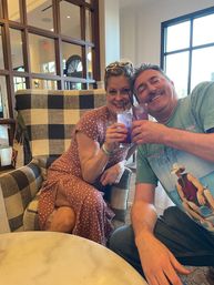 Two smiling people toasting purple iced cocktails with straws while seated in a black-and-white checkered armchair in a bright indoor lounge