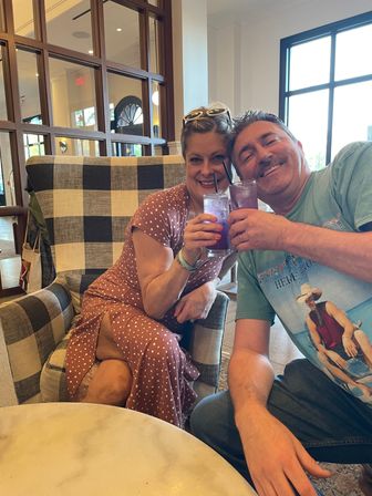 Two smiling people toasting purple iced cocktails with straws while seated in a black-and-white checkered armchair in a bright indoor lounge