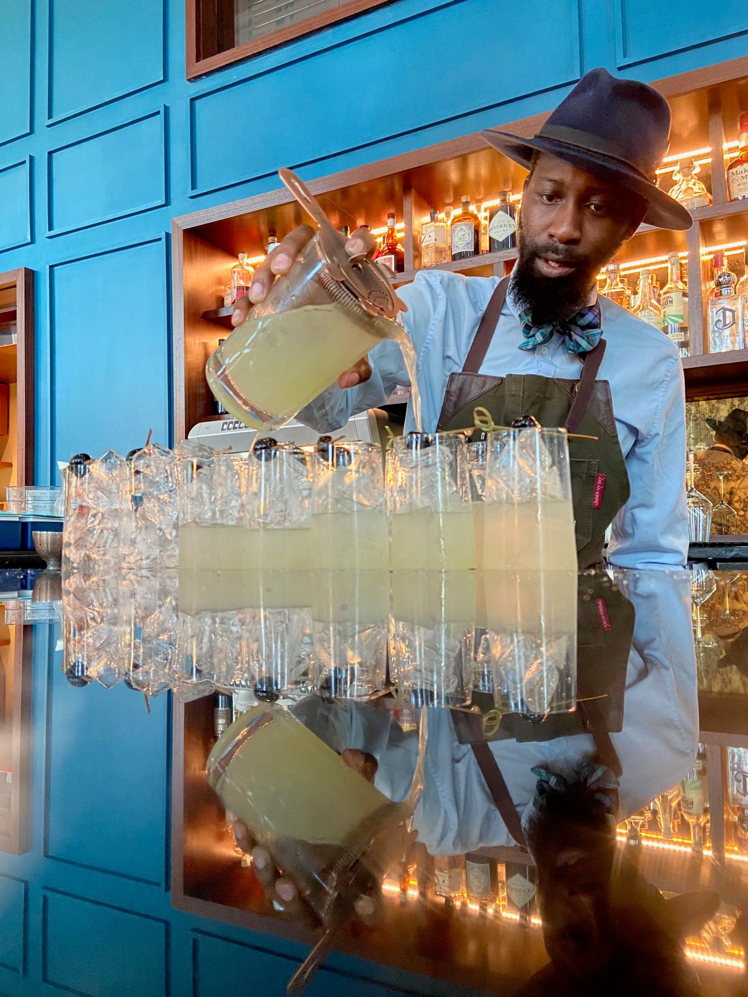 Mixologist wearing a hat and bow tie pouring pale citrus cocktails into a row of ice-filled highball glasses at a modern blue-paneled bar with a reflective countertop.