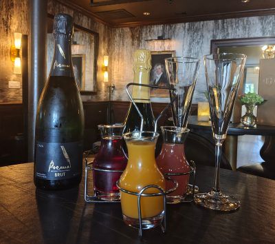 Sparkling wine bottle with three small glass carafes of orange, red, and purple juices and two champagne flutes arranged on a dark wooden bar table in a cozy rustic pub brunch setting.