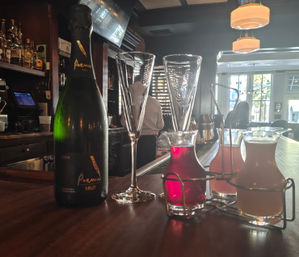 Sparkling Cava bottle and two champagne flutes on a wooden bar counter with three small glass carafes of colorful cocktail mixers in a metal rack, backlit cozy cocktail bar interior and bartender silhouette by sunlit windows.