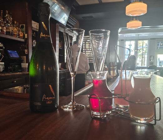 Sparkling Cava bottle and two champagne flutes on a wooden bar counter with three small glass carafes of colorful cocktail mixers in a metal rack, backlit cozy cocktail bar interior and bartender silhouette by sunlit windows.