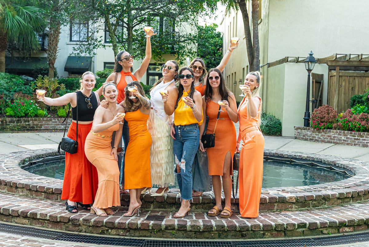 Group of friends in orange outfits toasting cocktails around a brick fountain in a sunny outdoor courtyard — festive brunch celebration