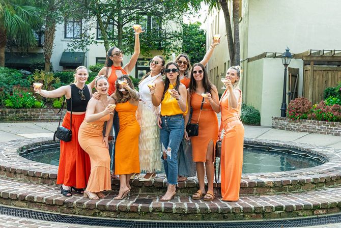 Group of friends in orange outfits toasting cocktails around a brick fountain in a sunny outdoor courtyard — festive brunch celebration