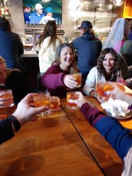 Friends cheering as they toast amber cocktails over a wooden table in a lively bar with a TV and beer taps behind them.
