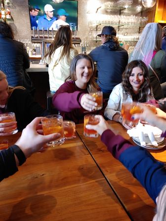 Friends cheering as they toast amber cocktails over a wooden table in a lively bar with a TV and beer taps behind them.