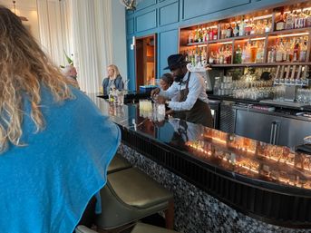 Upscale cocktail bar interior with bartender in a hat mixing drinks behind a glossy black counter reflecting warm-lit liquor shelves and seated patrons