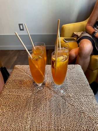 Four tall iced tea drinks with lemon slices and bamboo straws on a woven rattan table in a cozy lounge with a yellow armchair