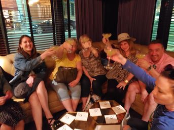 Group of friends toasting with margarita-style cocktails while seated on a curved leather sofa in a dim cocktail lounge, low tables with napkins in the foreground — lively night out.