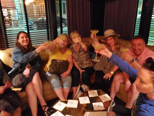 Group of friends toasting with margarita-style cocktails while seated on a curved leather sofa in a dim cocktail lounge, low tables with napkins in the foreground — lively night out.