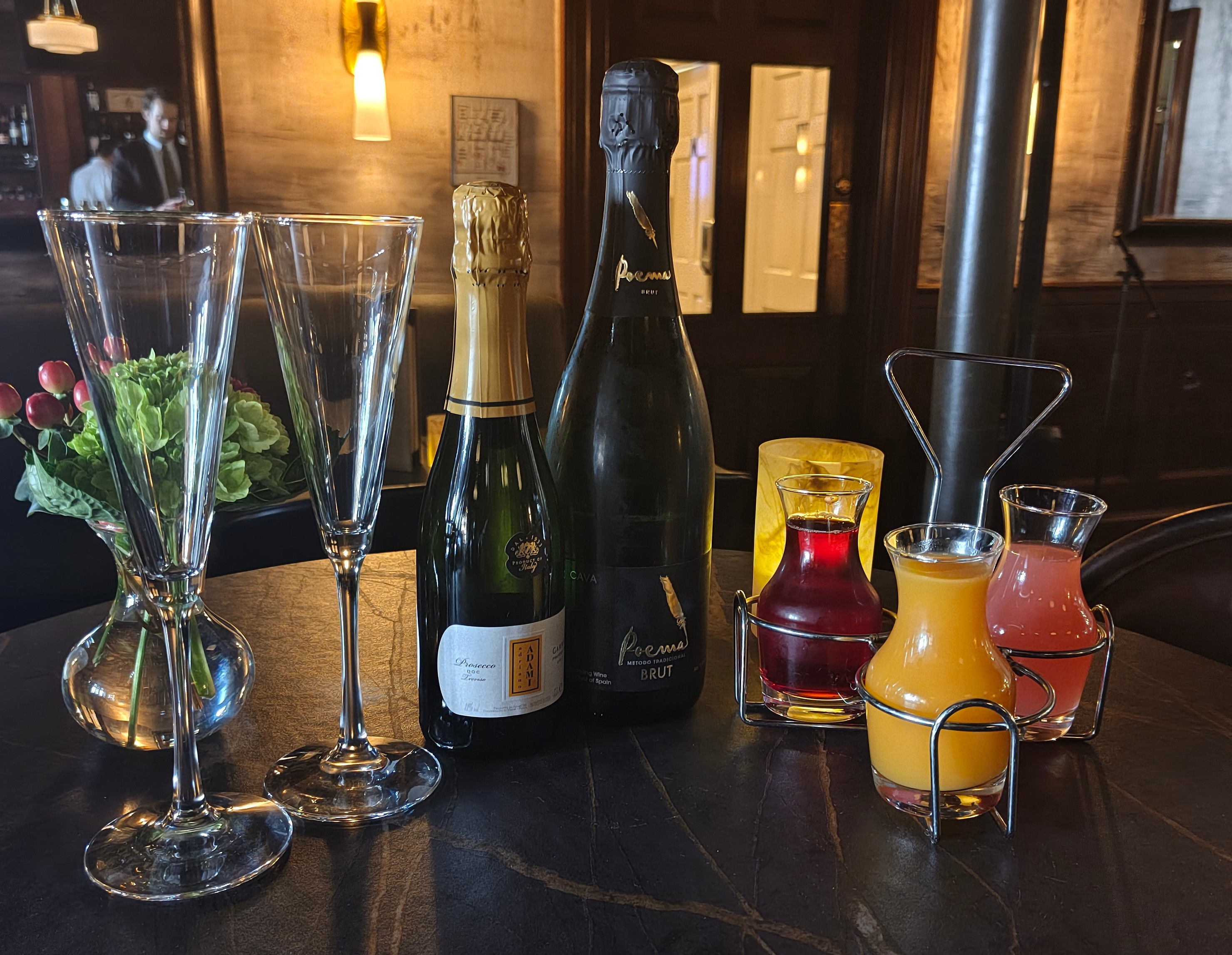 Upbeat bar scene: two champagne bottles and two flutes on a dark table at an upscale lounge, three colorful juice carafes in a metal rack, a small floral vase and warm ambient lighting — perfect brunch or celebration setup.