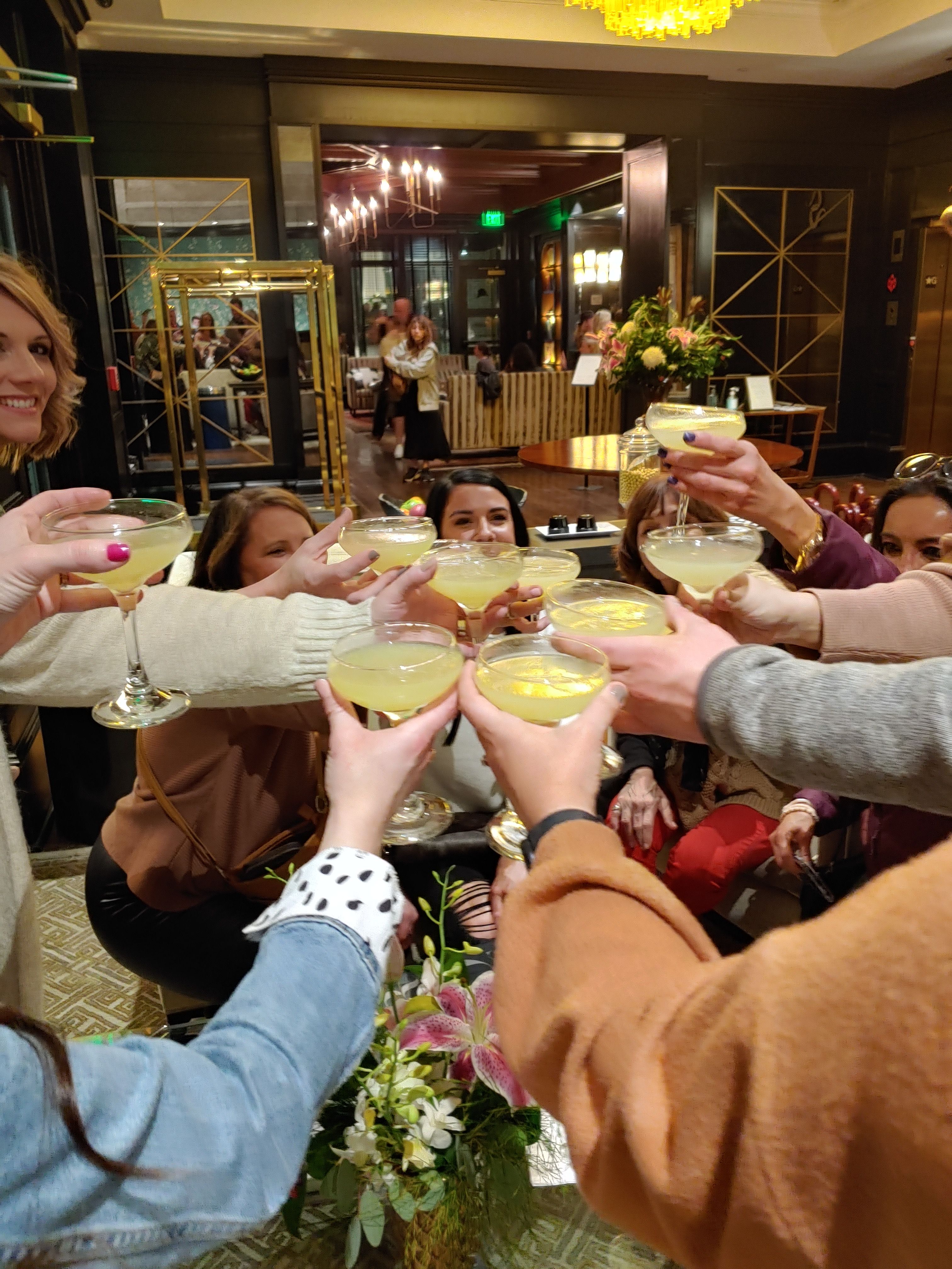 Friends toasting with lemon-yellow cocktails in coupe glasses in an upscale hotel lounge, floral centerpiece and warm chandelier-lit interior.