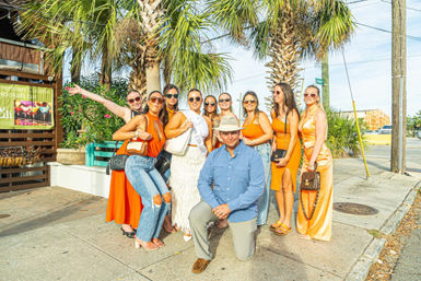 Sunny coastal street scene with a man in a blue shirt and hat kneeling and a group of women in orange dresses, denim and sunglasses posing together under palm trees with handbags on a sidewalk.
