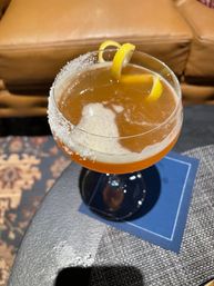 Amber cocktail in a coupe glass with a sugared rim and lemon twist, resting on a blue napkin on a side table in a cozy lounge setting
