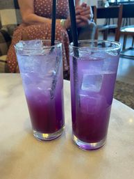 Vibrant purple iced drinks with ice and black straws in two tall clear glasses on a marble café table, blurred diner and chairs in the background