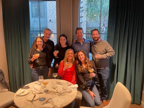 Seven adults smiling and toasting with cocktails around a round table in a cozy urban restaurant booth, green curtains and a window showing a brick building and palm tree outside.