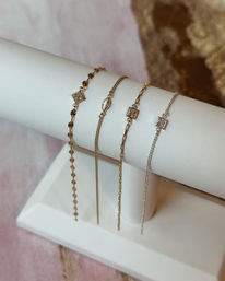Close-up of four delicate gold and silver bracelets on a white leather jewelry display — minimalist chains with geometric links and sparkling stone accents.