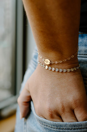 Close-up of a wrist wearing two delicate layered gold chain bracelets with small disc charms, hand tucked into a denim pocket — minimalist fashion jewelry detail
