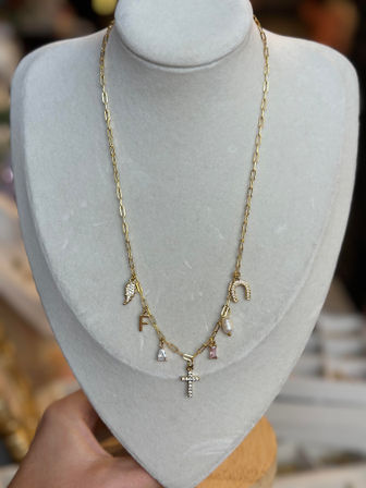 Gold paperclip-chain charm necklace with pavé cross pendant, horseshoe, freshwater pearl, pink and clear gemstone charms and an 'F' initial, shown on a gray velvet bust jewelry display.