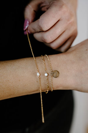 Close-up of a wrist and hand wearing three layered gold bracelets — a delicate chain with a small pearl, a chain with a clear gemstone, and a gold disc charm engraved 'MAMA' — minimalist jewelry detail shot.