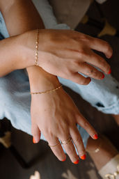 Close-up of hands resting on denim featuring bright orange nail polish and a small star finger tattoo, showcasing delicate gold chain bracelets and stacked gold rings for a casual jewelry look.