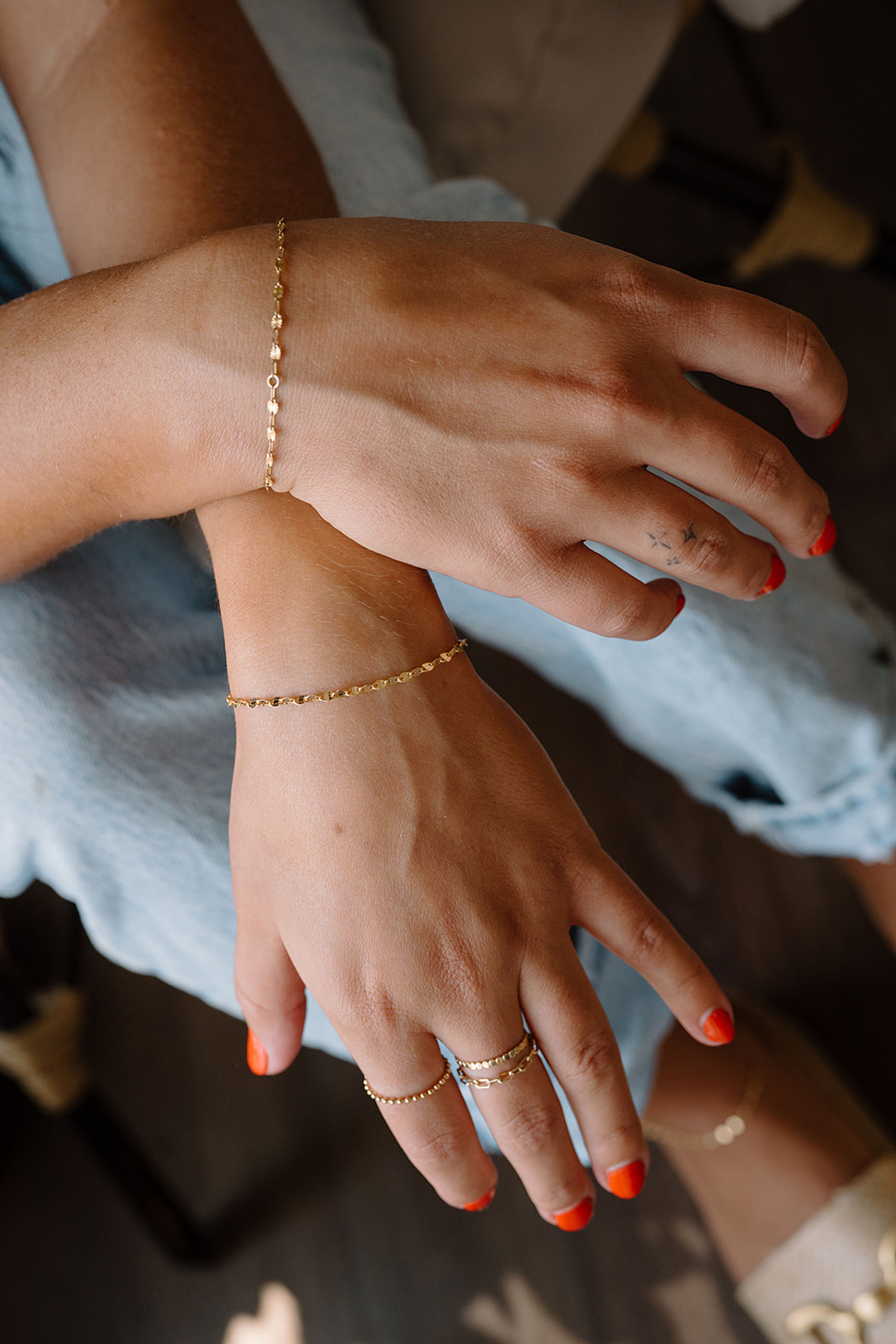 Close-up of hands resting on denim featuring bright orange nail polish and a small star finger tattoo, showcasing delicate gold chain bracelets and stacked gold rings for a casual jewelry look.