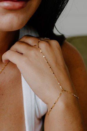 Close-up of woman's hand wearing a delicate gold hand-chain bracelet, minimalist jewelry styled with a white tank top in soft natural light