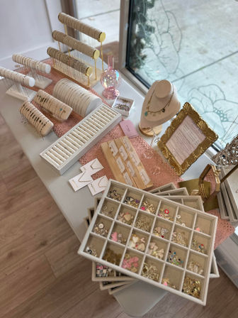 Boutique jewelry display by a window featuring cream and gold necklace bust, bracelets on rolled stands, organized charm and ring trays, and a pink lace runner on a white table.
