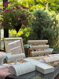 Outdoor patio jewelry display featuring delicate gold and silver bracelets and chain necklaces on white padded and gold tiered stands, framed price list, pink hanging basket flowers and lush garden greenery in the background.