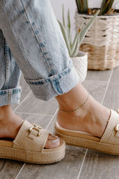 Close-up of cuffed light-wash jeans, green beaded anklet and woven beige platform slide sandals on gray tile floor with potted plants — casual boho summer style