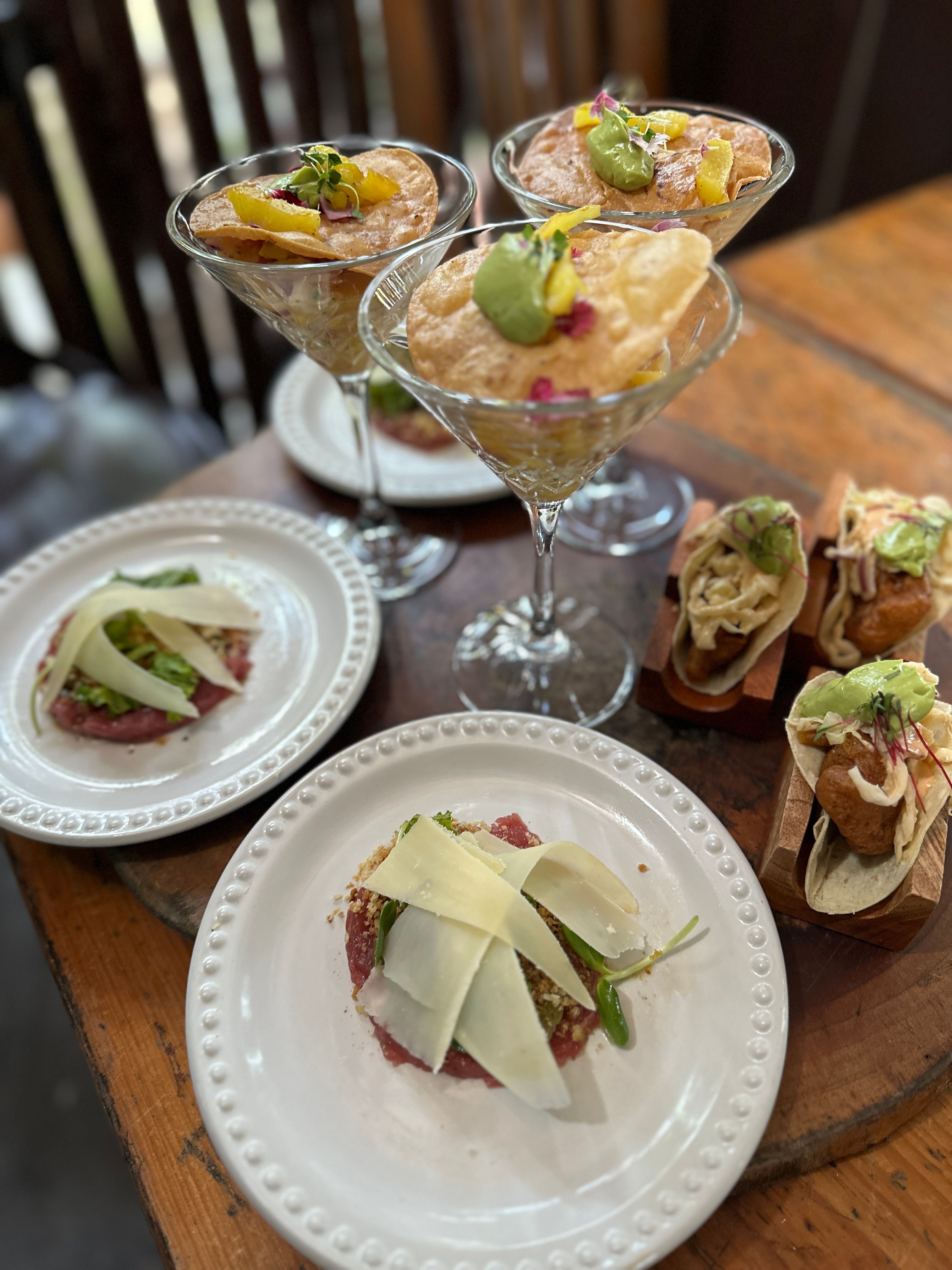 Gourmet appetizer spread on a wooden table: martini glasses filled with crispy tortilla chips topped with avocado crema and mango, two plates of tuna tartare finished with shaved Parmesan, and mini tacos in wooden holders — cozy restaurant small plates.