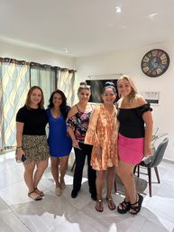 Five smiling women posing together in a bright modern living room, wearing colorful summer outfits; patterned curtains, wall clock and dining chairs visible in the background.