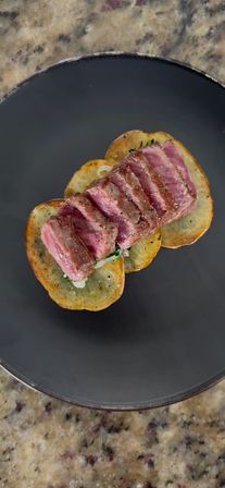 Top-down shot of sliced medium-rare steak served atop golden roasted potato rounds with herb garnish on a matte black plate, gourmet steak and potatoes plating.