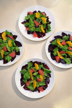 Top-down view of four white plates with a bright beet and citrus salad — roasted beet slices topped with arugula and grapefruit and orange segments on a light countertop.