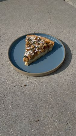 Sunlit slice of crumb-topped fruit pie dusted with powdered sugar and tiny herb sprigs on a blue ceramic plate set on a concrete patio