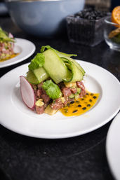 Close-up of gourmet tuna tartare with diced tuna and avocado, topped with curled cucumber ribbons, cilantro and a radish slice, finished with bright passionfruit sauce on a white plate in a restaurant setting.