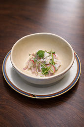 Gourmet seafood ceviche with thin radish slices, cilantro and microgreens in a beige bowl on a blue-and-gold rimmed plate, photographed on a dark wooden table.