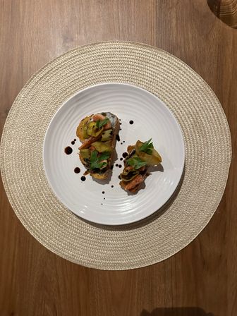 Two crostini topped with grilled zucchini, tomato, fresh parsley and balsamic glaze on a white plate set on a woven placemat over a wooden table