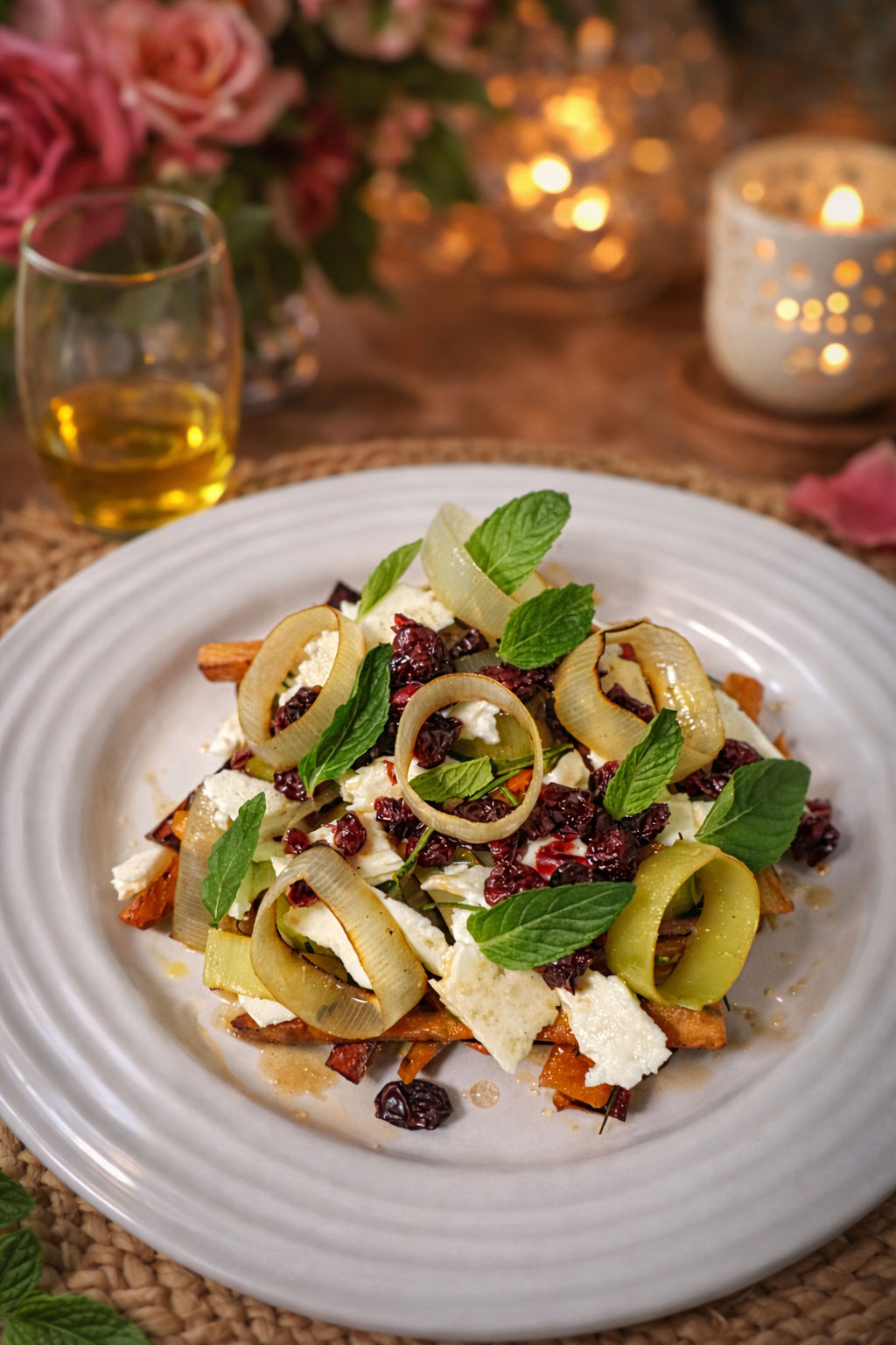 Gourmet roasted carrot and feta salad topped with pickled leek rings, dried cranberries and fresh mint leaves on a white plate in a cozy candlelit table setting