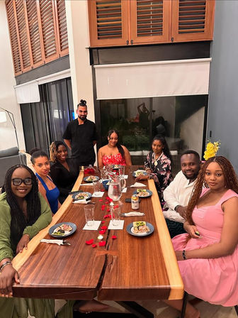 At-home dinner party: smiling guests and a chef around a large live-edge wooden table set with appetizers, rose petals and a water pitcher in a modern dining room with shuttered windows.