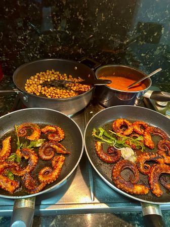 Sizzling Mediterranean-style grilled octopus tentacles in two frying pans with herbs and roasted garlic, alongside a skillet of spiced roasted chickpeas and a pot of red tomato sauce on a home stovetop.