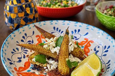 Roasted baby corn topped with crumbled cheese, fresh herbs and chili flakes, served with a lime wedge in a colorful Mexican-style ceramic bowl